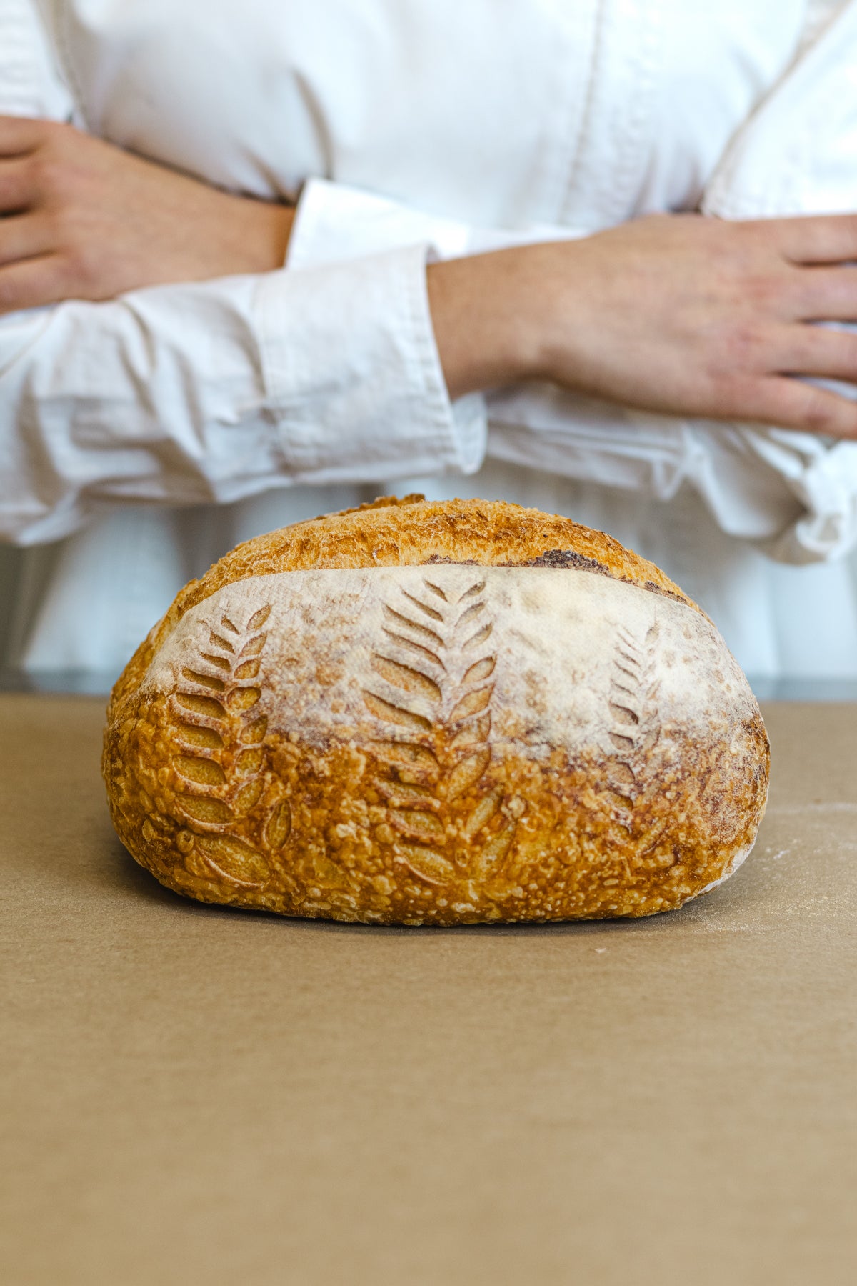 A rustic round artisan bread loaf with a flour-dusted crust and leaf imprint, sitting on a brown surface with a person in a white shirt blurred in the background.