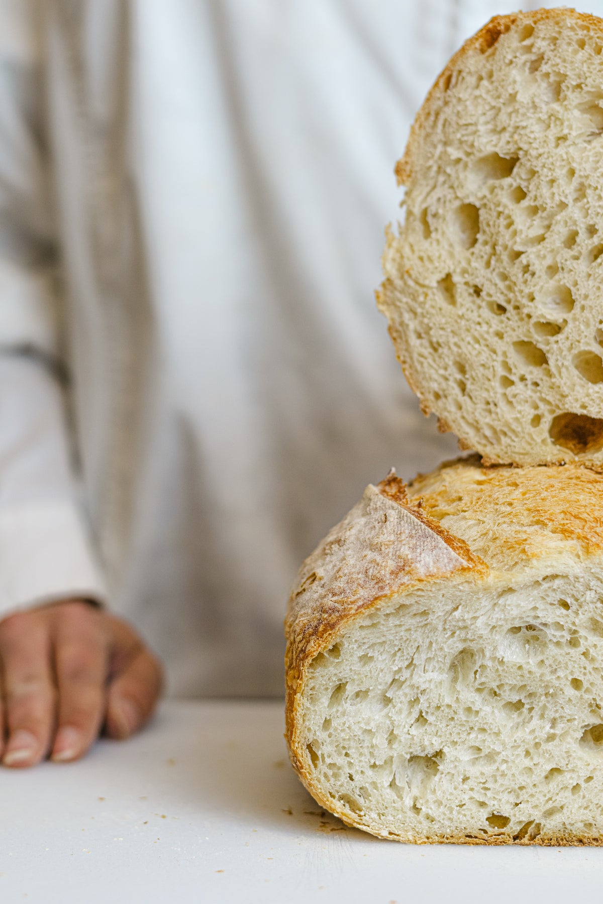 Close-up of a rustic round bread loaf cut into two halves, showing the airy crumb structure and golden-brown crust, with a person's hand and torso blurred in the background.