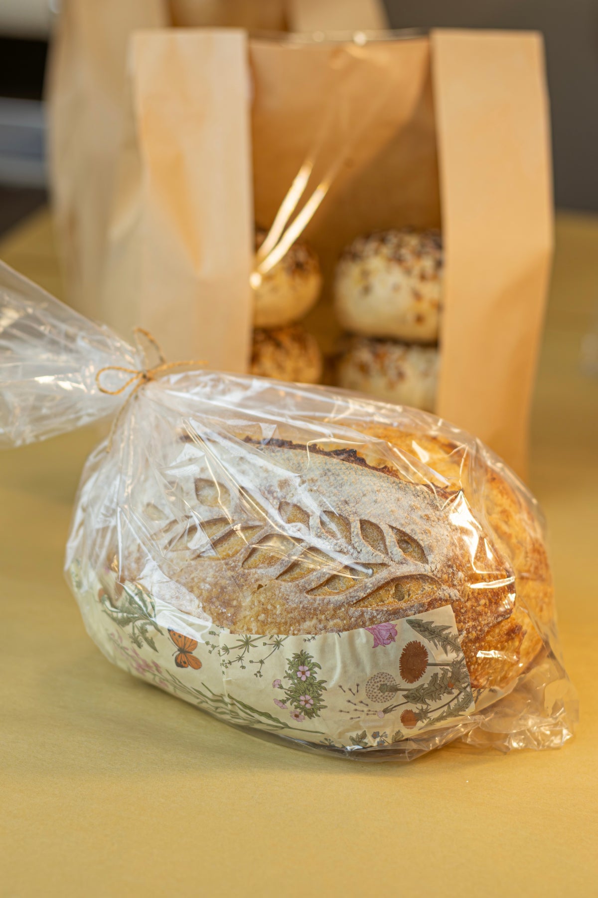 A loaf of rustic bread wrapped in clear cellophane and tied with a string, placed on a yellow surface with a brown paper bag of more bread behind it.