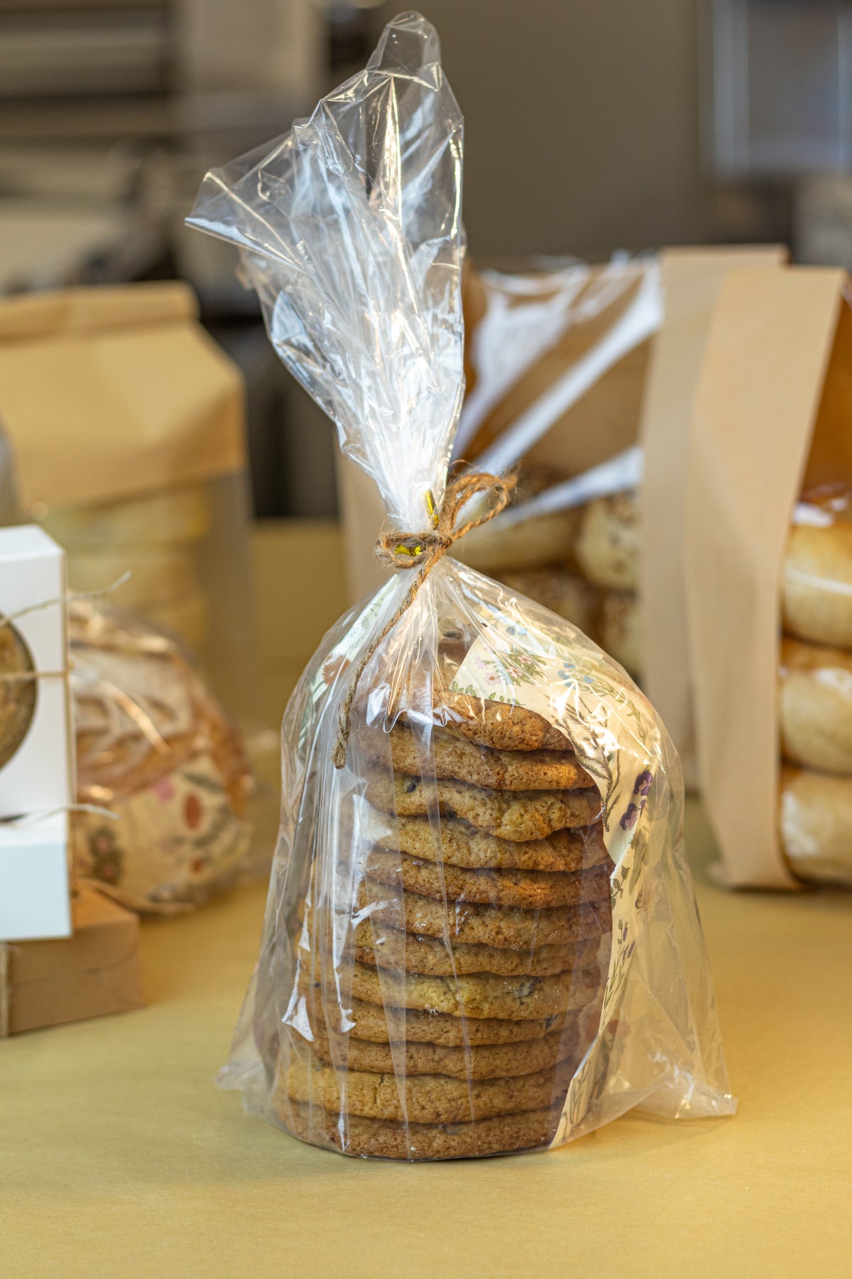 A clear cellophane bag tied with twine at the top, filled with a stack of round cookies, standing on a beige tabletop. Other packaged baked goods are blurred in the background.