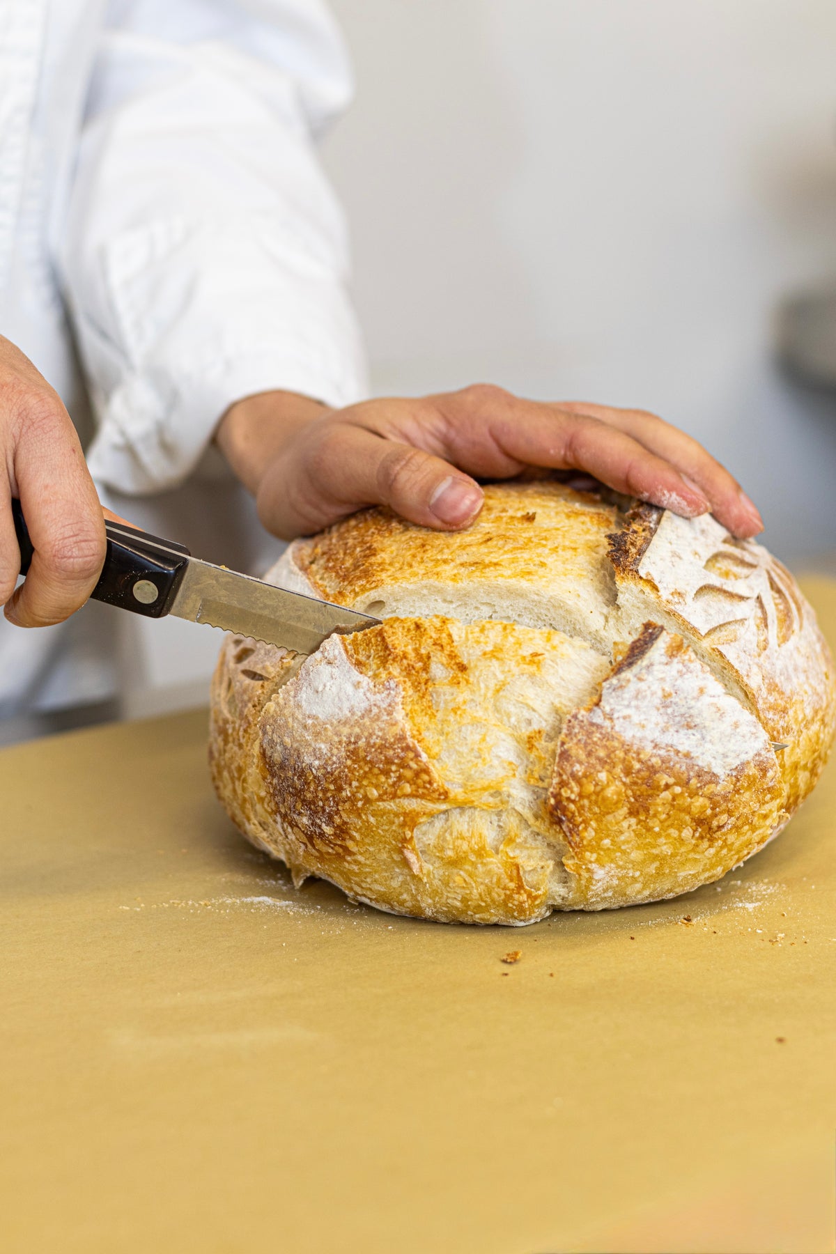 A close-up of a person in a white chef's coat slicing a freshly baked sourdough bread loaf on a wooden surface.