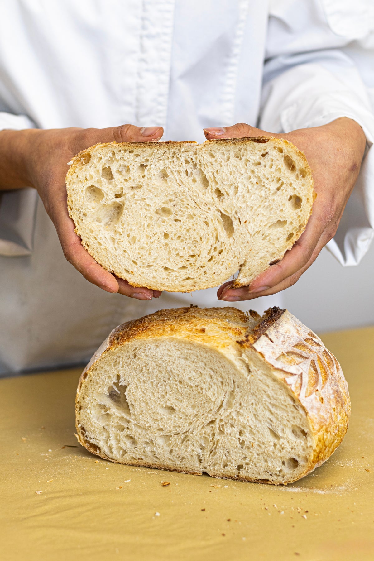 A person in a white chef's coat holds up a thick slice of rustic artisan bread to show its airy crumb structure, with a larger loaf of the same bread resting on a yellow surface below.