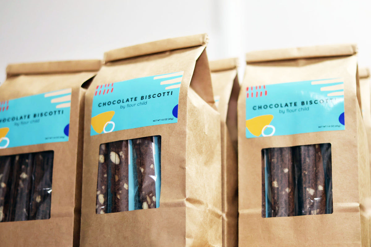 Three brown kraft paper bags of chocolate biscotti with light blue labels, arranged side by side against a plain light background.