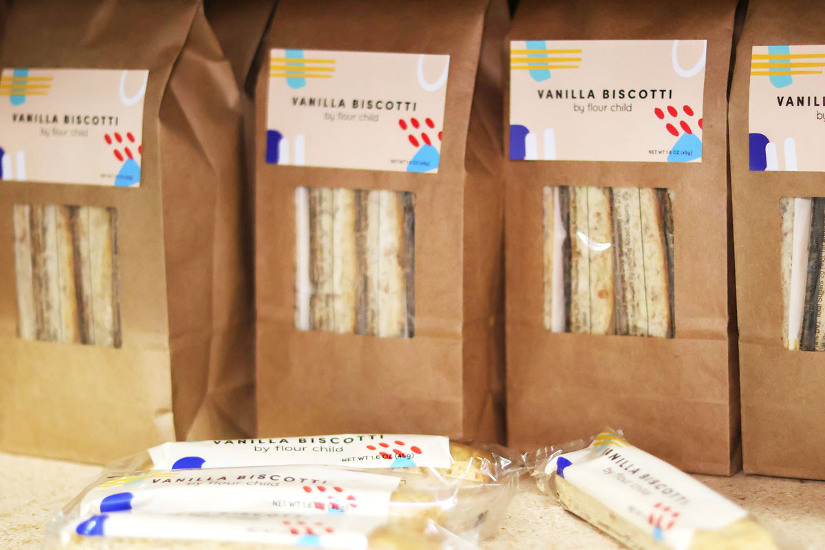 Several brown paper bags of vanilla biscotti cookies with clear windows are lined up, with a few individually wrapped biscotti in the foreground on a light surface.