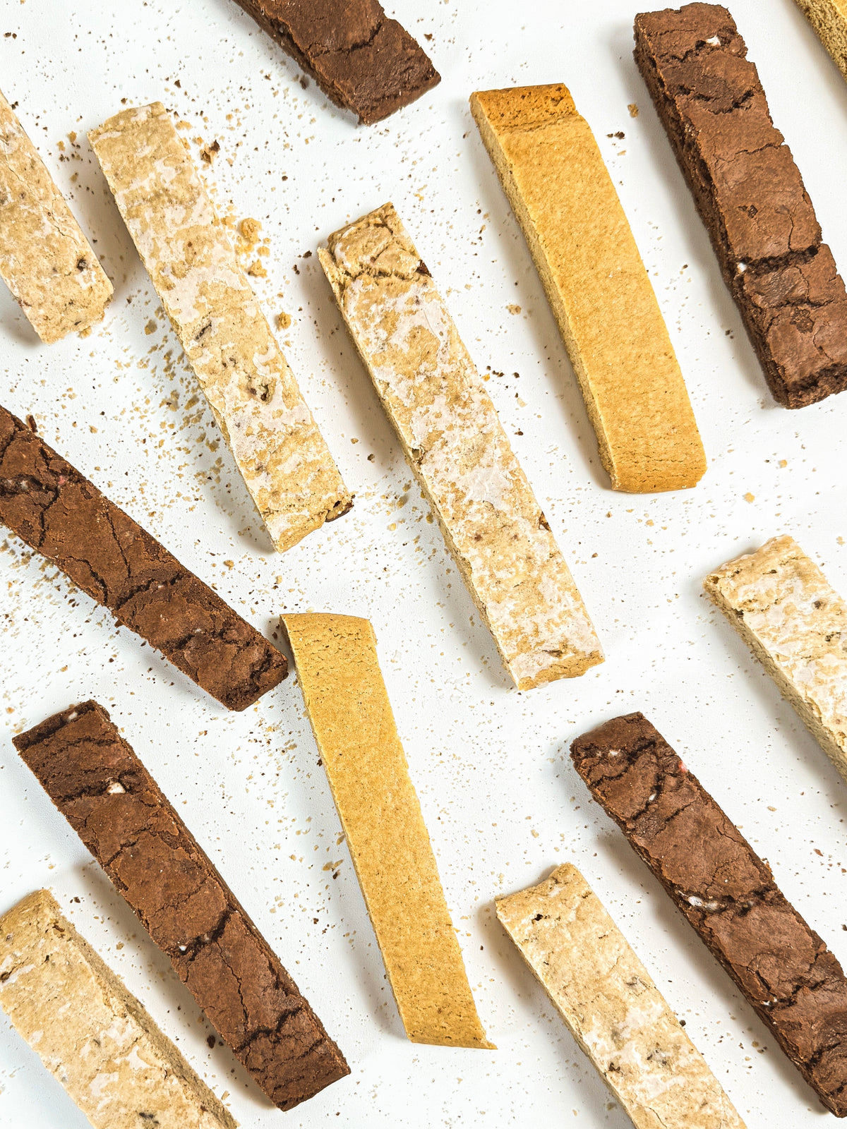 An overhead flat-lay of assorted biscotti sticks on a white background, featuring both dark chocolate brown and light golden-beige varieties.